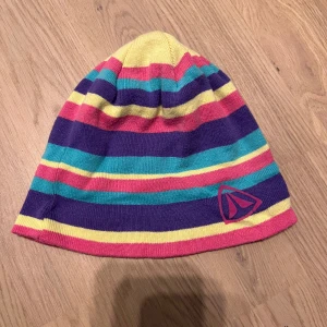 Randig mössa med färgglada ränder - Säljer en färgglad mössa med ränder i gult, rosa, lila och turkos. Mössan har en logotyp på sidan och är perfekt för att lysa upp vinterdagarna.