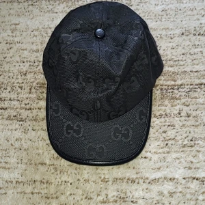 Svart jumbo keps från Gucci - Snygg svart keps från Gucci med diskret mönster av märkets logotyp. Kepsen har en justerbar rem baktill för perfekt passform och är tillverkad i ett slitstarkt material.