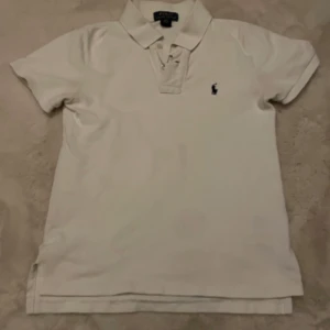 Vit kortärmad pikétröja från Ralph Lauren - Säljer en klassisk vit pikétröja från Ralph Lauren med korta ärmar och en liten broderad logga på bröstet. Perfekt för en stilren och avslappnad look. Tröjan har en krage och knappar framtill.