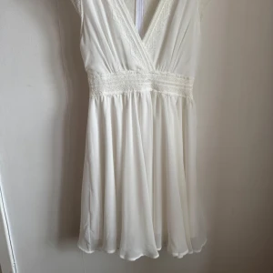 Vit klänning från TFNC London - Säljer en vacker vit klänning från TFNC London med spetsdetaljer och omlott-design. Klänningen har en v-ringad hals och är ärmlös, perfekt för sommarens festligheter. Den har en luftig kjol som ger en fin siluett.