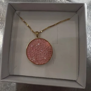 Halsband med rosa hänge från Caroline Svedbom - Helt nytt halsband! Aldrig använt Elegant halsband med ett runt rosa hänge infattat i guld. Hänget har en glittrig yta som fångar ljuset vackert. Perfekt för att ge en färgklick till din outfit.