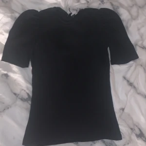 Svart topp från Pieces - Säljer en stilren svart topp från Pieces. Toppen har korta ärmar med en lätt puffig design och är tillverkad i ett mjukt material. Perfekt för att matcha med olika outfits. Hör av dig vid intresse!