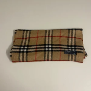 Rutig halsduk från Burberry - Säljer en klassisk rutig halsduk från Burberry i 100% lammull. Den har en beige bas med svarta, vita och röda rutor. Perfekt för att hålla dig varm och stilren under kyliga dagar. Tillverkad i England. Nypris 1999 mitt pris 569