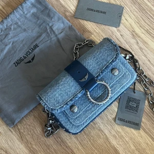 Blå jeansväska från Zadig & Voltaire - Helt ny snygg  blå jeansväska från Zadig & Voltaire med en cool kedjerem och metallringdetalj på framsidan. Väskan har en praktisk magnetknäppning och en stilren design som passar perfekt för en trendig look. Perfekt för att ge din outfit en edgy touch!