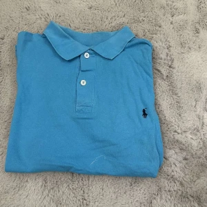 Blå pikétröja från Ralph Lauren - Säljer en klassisk blå pikétröja från Ralph Lauren med krage och knappar framtill. Perfekt för en stilren och avslappnad look. Den har en liten broderad logga på bröstet.