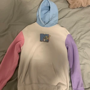 MTV hoodie med färgglada ärmar - Säljer en cool MTV hoodie med en vit bas och färgglada ärmar i rosa och lila. Huvan är ljusblå och tröjan har en stor MTV-logga på framsidan. Perfekt för dig som vill ha en färgglad och bekväm stil!