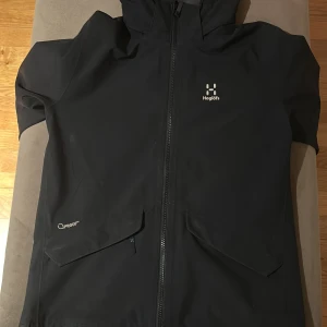 Svart regnjacka från Haglöfs - Säljer en svart regnjacka från Haglöfs med dragkedja och huva. Jackan har en stilren design med reflekterande detaljer och är tillverkad i ett vattentätt material. Perfekt för regniga dagar! Passar också 160 cm 