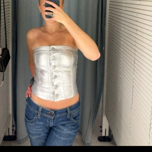 Silverfärgad korsett från Amisu - Säljer en snygg silverfärgad korsett från Amisu. Den är axelbandslös och har knappar framtill. Perfekt för att ge en stilren look med en touch av glamour. Passar bra till jeans eller kjol för en chic outfit.