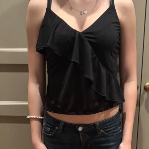 Svart linnetopp med volangdetalj - Snygg svart linnetopp med smala axelband och en elegant volangdetalj framtill. Perfekt för en stilren look.