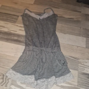 Grå jumpsuit med spetsdetaljer - Säljer en söt grå jumpsuit med vita spetsdetaljer vid halsringning och ben. Den har smala axelband och en knytning i midjan för en bekväm passform. Perfekt för en avslappnad dag hemma eller sommarens varma kvällar.