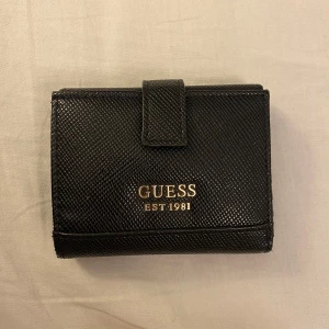 GUESS plånbok - Snygg svart plånbok från Guess med guldfärgad dragkedja. Plånboken har flera kortfack och en praktisk dragkedjeficka för mynt. Perfekt för att hålla ordning på dina kort och kontanter.