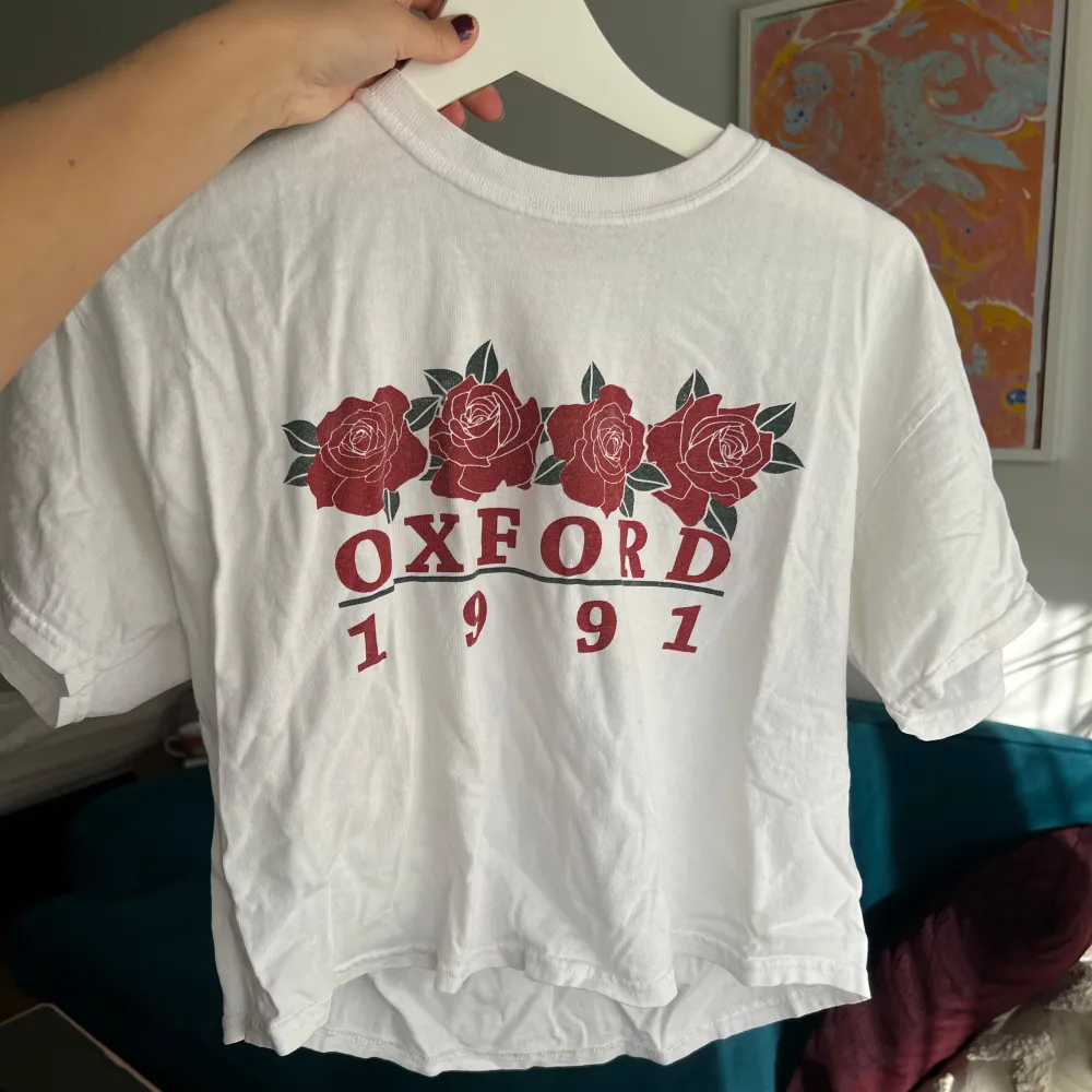 Säljer en vit t-shirt med tryck av röda rosor och texten 'Oxford 1991'. T-shirten har en croppad stil och korta ärmar, perfekt för en avslappnad look. Passar mellan xs-m, bra skick men lite skrynklig . T-paidat.