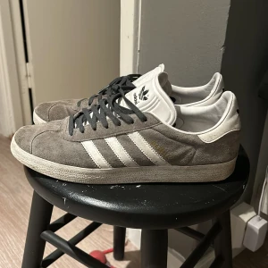 Grå Adidas Gazelle sneakers - Säljer ett par klassiska grå Adidas Gazelle sneakers med vita detaljer och snörning. Skorna har en stilren design med tre vita ränder på sidan och en bekväm passform. Perfekta för en avslappnad stil. Pris kan såklart diskuteras!