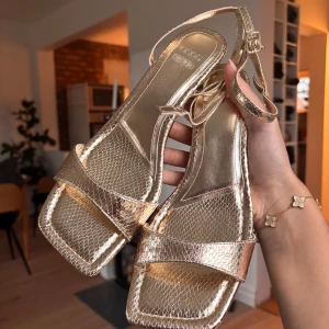 Guldiga sandaler med klack - Snygga guldiga sandaler med en elegant klack och smala remmar. Perfekta för att ge en glittrig touch till din outfit. De har en öppen tå och justerbar rem runt ankeln. Helt oanvända och kommer i sin förpackning. 