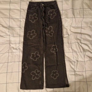 Grå blommiga jeansbyxor från H&M - Snygga grå jeansbyxor från H&M med ett coolt blommönster. Byxorna har en hög midja och en wide fit, vilket ger en avslappnad look. De har slitsar vid bensluten för extra stilpoäng. Perfekta för att ge din outfit en unik touch!