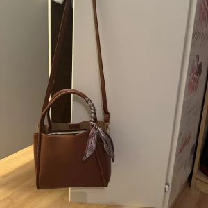 Brun axelväska med randig detalj - Snygg brun axelväska i skinn med en randig knytrosett på handtaget. Väskan har en praktisk dragkedja och en justerbar axelrem. Använd endast två gånger. Lagom stor, får plats med hyfsat mycket. Köptes för 500