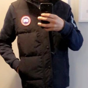 Svart Canada Goose väst - Snygg svart dunväst med en tydlig logotyp på bröstet. Perfekt för kyligare dagar och ger en stilren look. Västen har en dragkedja framtill och är designad för att hålla dig varm och bekväm.