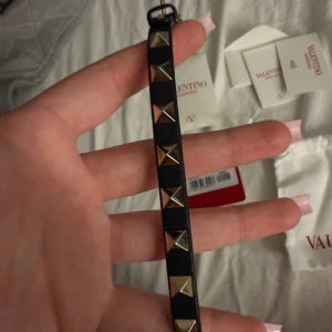 Svart armband med nitar från Valentino - Snyggt svart armband från Valentino med guldiga nitar. Armbandet har en justerbar rem och kommer med originalförpackning och påse. Perfekt för att ge en najs touch till din stil. Nypris 2950kr, Köpt på NK i Göteborg!