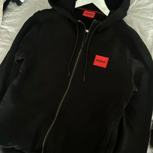 Svart hoodie från Hugo - Säljer en stilren svart hoodie från Hugo med röd logga på bröstet. Den har dragkedja och huva med snörning. Perfekt för en casual look.