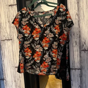 Blommig blus med korta ärmar - Säljer en blommig blus med korta ärmar i svart, orange och grått. Blusen har en v-ringad hals och ett färgglatt blommönster som ger en somrig känsla. Perfekt för att liva upp garderoben!