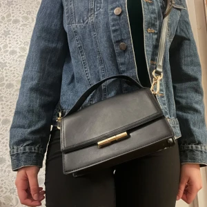 Svart axelväska med gulddetalj - Snygg svart axelväska i skinn med en gulddetalj på framsidan. 