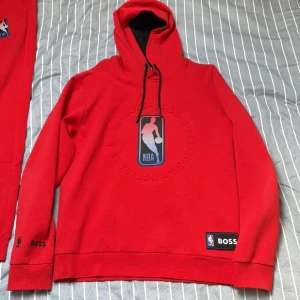 Röd NBA x BOSS hoodie + byxa - Riktigt snygg röd hoodie från BOSS i samarbete med NBA. Stor broderad NBA-logga framtill, svarta snören i huvan och svart innerfoder. BOSS- och NBA-märke på ärmen och nertill. Perfekt för dig som gillar basket och streetwear.