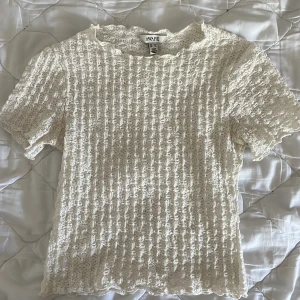 Vit spetsblus från Aware - Superfin vit blus från Aware med korta ärmar och vacker spetsstruktur. Blusen har en lätt vågig kant vid halsen och är tillverkad i ett genombrutet spetsmaterial som ger en romantisk vibe. Perfekt att styla med jeans eller kjol för en söt look.