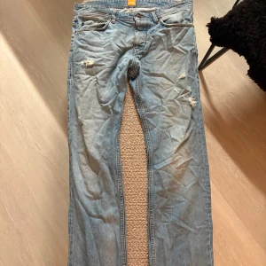 Boss Orange ljusblå jeans med slitningar - Säljer ett par ljusblå jeans från Boss Orange med snygga slitningar på låren och klassiska femficksdetaljer. Jeansen har raka ben och är tillverkade i mjukt denimtyg. Perfekta för dig som gillar en avslappnad och trendig look.