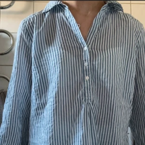 Blåvit randig henley skjorta - Snygg blåvit randig henley skjorta med klassisk krage och knappar framtill. Skjortan har lång ärm och är tillverkad i ett lätt och skönt bomullstyg, perfekt för en avslappnad och stilren look.