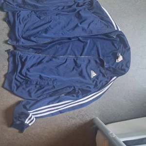 Marinblå Adidas vindjacka med stripes - Säljer en marinblå vindjacka från Adidas med klassiska vita stripes längs ärmarna och dragkedja framtill. Jackan har ribbade muddar, två fickor och Adidas-logga på bröstet. Perfekt för dig som gillar sportig och stilren look.