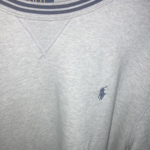 Grå sweatshirt från Polo Ralph Lauren XL - Klassisk ljusgrå sweatshirt från Polo Ralph Lauren i storlek XL. Tröjan har rund hals med randig ribbad kant och den ikoniska blå broderade loggan på bröstet. Perfekt basic med relaxed fit och mjukt bomullsmaterial.