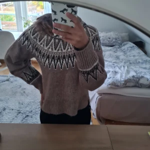 Beige stickad tröja med mönster - Säljer en beige stickad tröja med svart och vitt grafiskt mönster runt halsen och på ärmarna. Tröjan har ribbade muddar, hög hals och slits i nederkanten. Perfekt för kalla dagar och riktigt mysig att ha på sig. Endast använd fåtal gånger