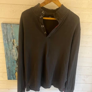 Grön långärmad tröja från BOSS - Säljer en stilren grön långärmad tröja från BOSS med ribbad hög krage och fyra knappar framtill. Tröjan är i mjuk bomull och har en klassisk passform som funkar till många outfits. 