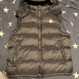Svart dunväst från Moncler - Säljer en svart dunväst från Moncler med huva och dragkedja framtill. Västen har två fickor med dragkedja även en inuti  och klassisk Moncler-logga på bröstet. Yttermaterialet är glansigt och quiltat, perfekt för lager-på-lager under kyliga dagar.riktigt skön till våren eller höst då den håller värmen. NFC Scan självklart och inga defekter överhuvudtaget . Priset är inte hugget i sten och byten kan diskuteras 