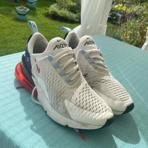 Nike Air 270 vita sneakers strl 40 - Nike Air 270 sneakers i vitt med blå och röda detaljer. Skorna har en luftig mesh-ovandel, stor röd air-bubbla i hälen och snörning med ljusblå öglor. Perfekta för dig som gillar sportig och modern stil. Snygg design och bekväm passform.