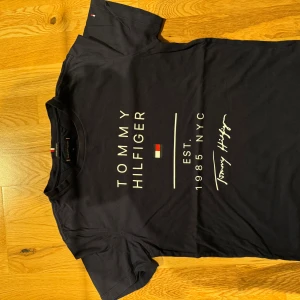 Mörkblå Tommy Hilfiger t-shirt - Snygg mörkblå t-shirt från Tommy Hilfiger med tryck framtill och klassisk logga. Tillverkad i ekologisk bomull, kortärmad och med rund hals. Perfekt för dig som gillar stilrena och ikoniska plagg med streetkänsla.