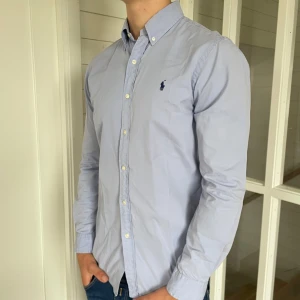 Polo Ralph lauren skjorta - Säljer denna riktigt snygga ljusblåa Ralph lauren skjortan! Den är i överlag mycket bra skick men har lite missfärgning i armhålorna, storlek M modell slim fit, den är 76 cm lång och 50 cm bred under armhålorna, jag på bilderna är 184 cm och väger 82 kg, hör gärna av dig vid frågor!