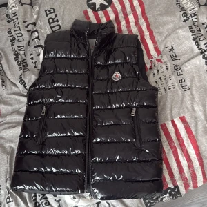Svart dunväst från Moncler - Snygg svart dunväst från Moncler med glansig finish och klassisk logga på bröstet. Västen har ståkrage, två dragkedjeförsedda fickor och quiltad design. Perfekt att slänga över en hoodie för extra värme. Stilsäker och tidlös look.