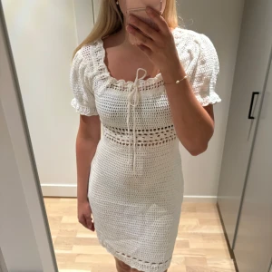 Vit virkad kortklänning med puffärm - Superfin virkad kortklänning i offwhite med puffärmar och söt snörning vid halsen. Klänningen har ett luftigt mönster i midjan och längs nederkanten, perfekt för varma dagar. Den är stickad i bomull och har en romantisk vibe.