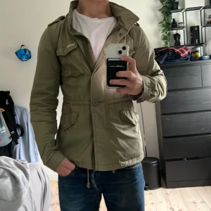 Field jacka Tommy hilfiger - Säljer en olivgrön field jacka från Hilfiger Denim med dragkedja och knappar framtill. Jackan har flera fickor, axelklaffar och justerbar midja med snörning. Perfekt för vår och höst. En defekt är att knappen har ramlat av men dragkedja funkar fortfarande, därav priset.