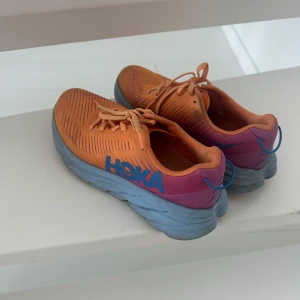 Orangea Hokka  sneakers med blå sula - Färgstarka Hokka sneakers i orange mesh med blå och lila detaljer samt blå tjock sula. Skorna har snörning och är perfekta för löpning eller träning. Snygg kontrast mellan färgerna och sportig design som sticker ut.