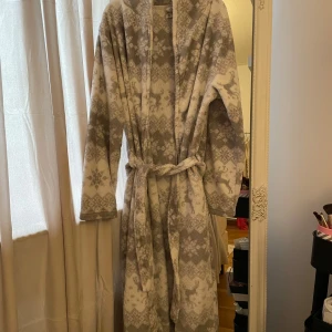 Mjuk grå morgonrock med vintermönster - Supermjuk och lång morgonrock i grått och vitt med klassiskt vintermönster av snöflingor och renar. Den har långa ärmar, sjalkrage och knytskärp i midjan. Perfekt för kalla dagar när du vill mysa hemma. KOM GÄRNA MED EGNA PRISFÖRSLAG!