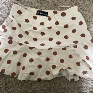 Vit prickig kjol från Zara - Supersöt kort kjol från Zara i vit med bruna prickar. Kjolen har en lätt volang nedtill med inbyggda shorts. 