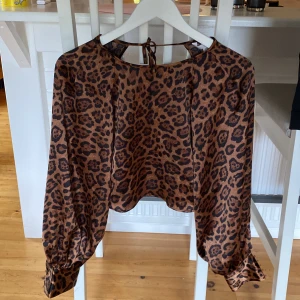 Leopardmönstrad blus med knyt - Säljer en brun och svart leopardmönstrad blus med långa puffärmar och snygga knappar vid ärmsluten. Blusen har en rund halsringning med öppning och knyt i nacken. Materialet är glansigt och känns lyxigt, perfekt för dig som gillar att sticka ut.