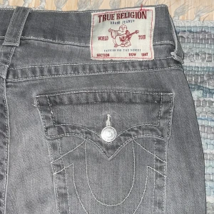 True Religion Ricky  - Säljer ett par grå jeans från True Religion, modell Ricky Relaxed Straight. De har klassiska kontrastsömmar, stora fickor med lock och silverfärgade knappar. Snyggt broderad logga på bakfickorna och patch med Buddha på linningen. Perfekt för dig som gillar street och premium denim.