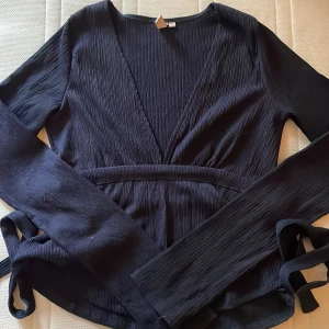 Marinblå topp med knytband - Säljer en marinblå topp från Nelly med långa ärmar och knytband i midjan, som man knyter i ryggen. Toppen har en djup v-ringning och är gjord i ett lätt, ribbat tyg som ger en snygg struktur. Perfekt för dig som gillar stilrena och trendiga plagg.