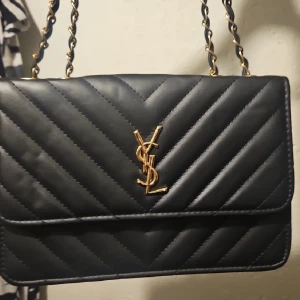 Svart quiltad YSL axelväska i skinn - Säljer en svart quiltad axelväska från YSL med klassisk kedjerem i guld och ikonisk YSL-logga framtill. Väskan är tillverkad i skinn och har ett stilrent, lyxigt uttryck som lyfter vilken outfit som helst.
