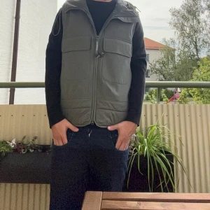 Grön dunvest från C.P. Company - Säljer en grön dunväst från C.P. Company med ikonisk huva med glasögondetaljer. Västen har två stora bröstfickor, dragkedja framtill och quiltad baksida. Tillverkad i mjukt syntetmaterial med stretch och återvunnen nylon. Perfekt för lager-på-lager och streetwear.