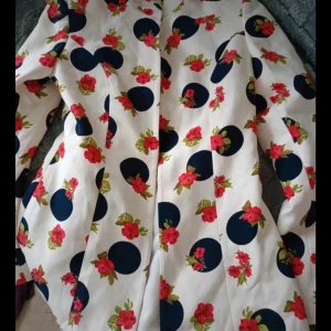 Vit blommig kappa med mörkblå prickar - Snygg kappa i vitt med stora mörkblå prickar och röda blommor över hela plagget. Långärmad och rak modell som ger en cool statement-look. Perfekt för dig som vill sticka ut med färg och mönster. Materialet känns slätt och lätt.