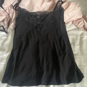 Svart linnetopp med spets från AMISU - Svart linnetopp från AMISU med tunna axelband och v-ringad halsringning dekorerad med snygg spets. Perfekt att styla lager på lager eller bära som den är. Mjuk och skön känsla med en feminin touch tack vare spetsdetaljerna.
